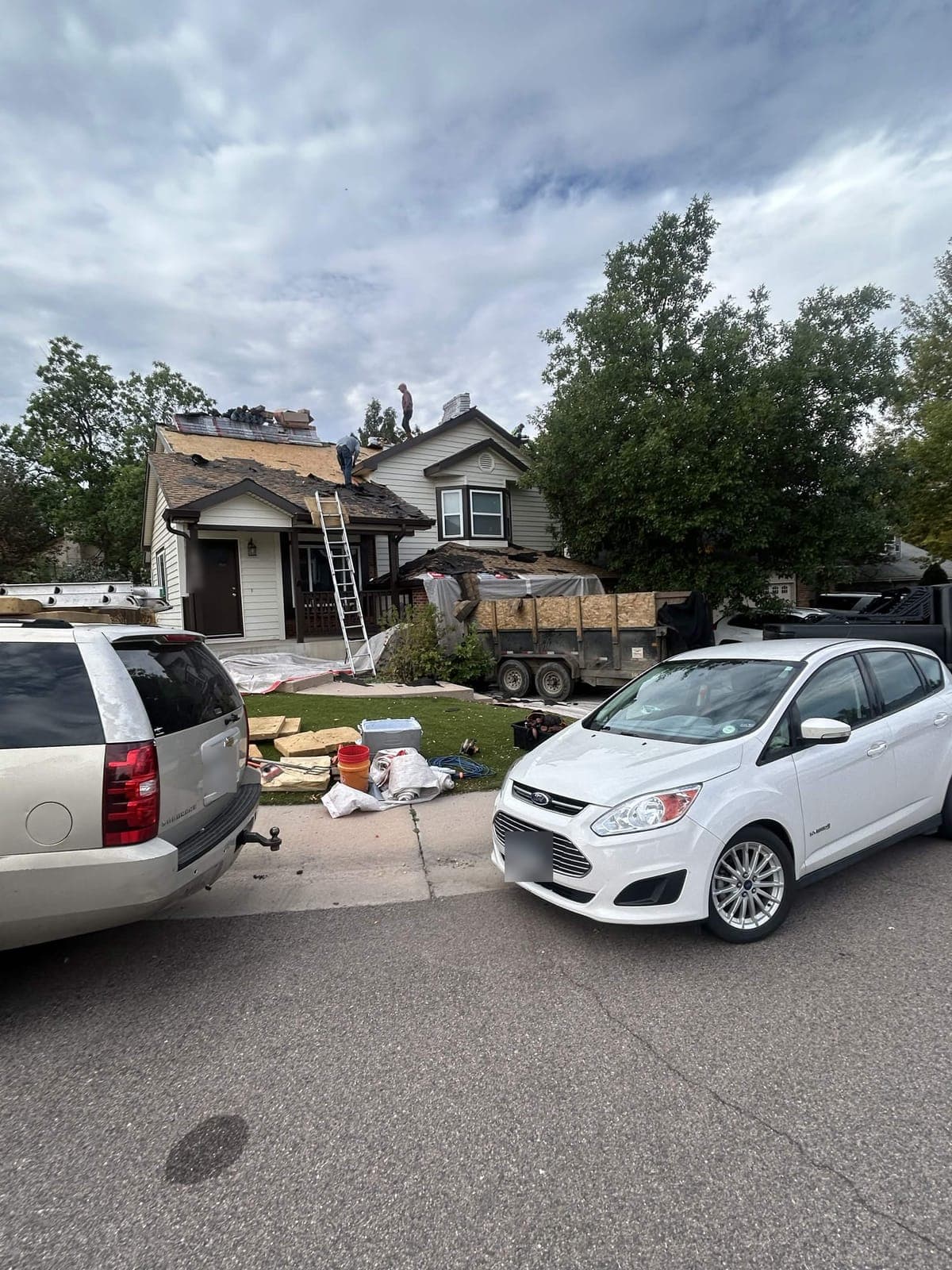Roofers tearing off old shingles on a two-story house, with a ladder at the front porch and a dump trailer of debris in the yard